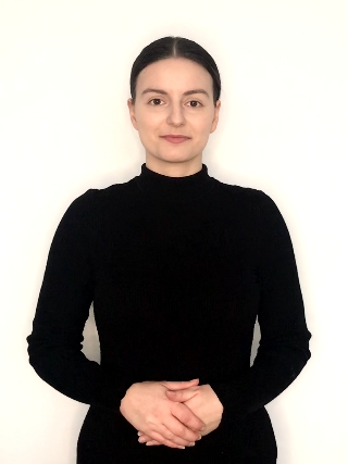 Kobieta stojąca na białym tle, ubrana w czarną bluzkę z długimi rękawami. Patrzy prosto w obiektyw, trzyma dłonie złożone przed sobą